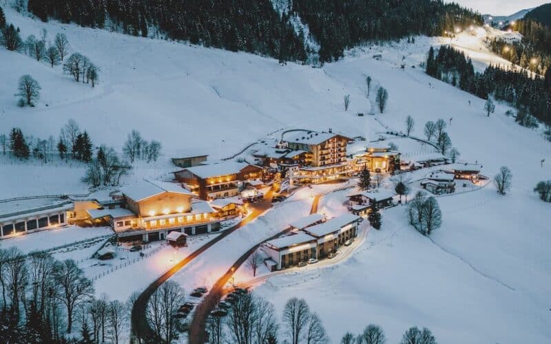 Luftaufnahme eines schneebedeckten Bergdorfs in der Abenddämmerung mit warm beleuchteten Gebäuden, umgeben von Bäumen und Hügeln.