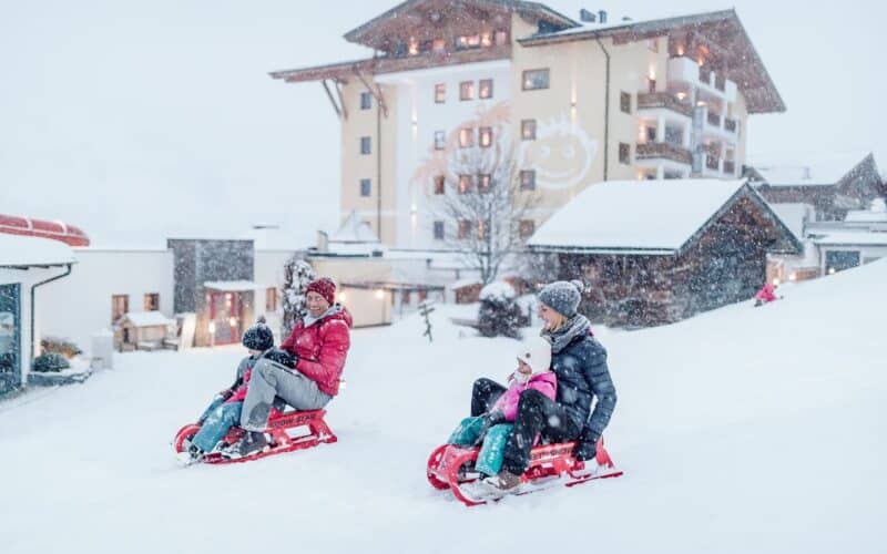 Zwei Erwachsene und zwei Kinder fahren bei Schneefall mit roten Schlitten einen verschneiten Hang vor einem Gebäude hinunter.