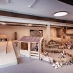 Die Kinder spielen in einem Innenbereich mit Holztiergehegen, kleinen Häusern und einer Bowlingbahn, an der auch ein Kind im Rollstuhl teilnimmt.