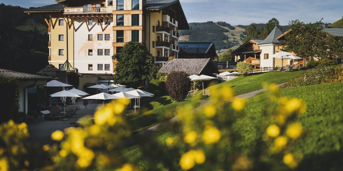 Ein mehrstöckiges gelbes Gebäude mit Balkonen und Sonnenschirmen im Freien, umgeben von grünen Hügeln und gelben Blumen im Vordergrund.