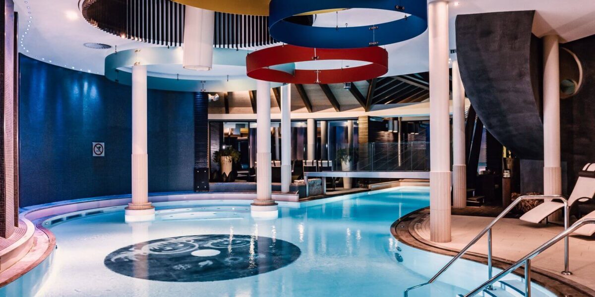 Hallenbad mit farbenfrohen, kreisförmigen Deckendekorationen, weißen Säulen, Stufen am Beckenrand und Liegestühlen, beleuchtet von Umgebungslicht.