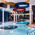 Hallenbad mit farbenfrohen, kreisförmigen Deckendekorationen, weißen Säulen, Stufen am Beckenrand und Liegestühlen, beleuchtet von Umgebungslicht.