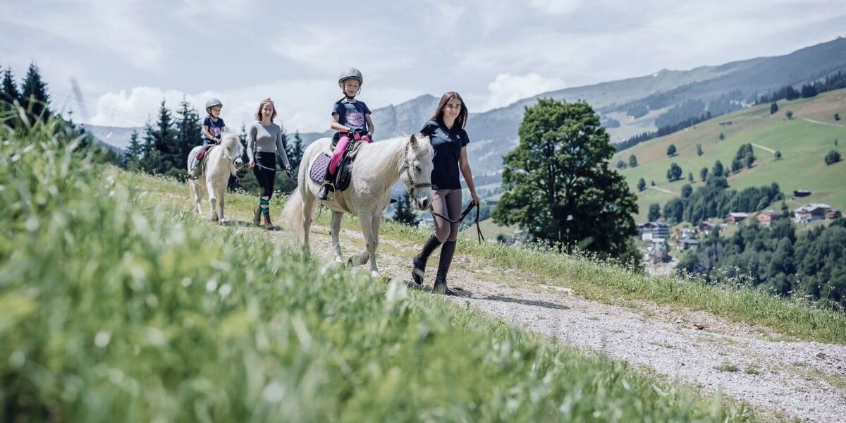 Drei Kinder reiten auf weißen Ponys einen Bergpfad entlang, wobei jedes von einem Erwachsenen zu Fuß geführt wird. Im Hintergrund sind Berge und ein Dorf unter einem teilweise bewölkten Himmel zu sehen.