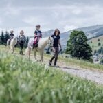 Drei Kinder reiten auf weißen Ponys einen Bergpfad entlang, wobei jedes von einem Erwachsenen zu Fuß geführt wird. Im Hintergrund sind Berge und ein Dorf unter einem teilweise bewölkten Himmel zu sehen.