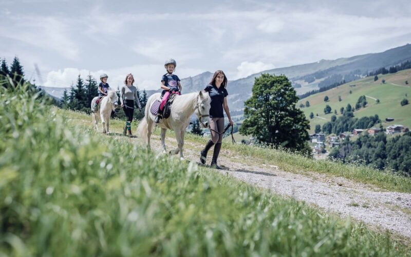 Drei Kinder reiten auf weißen Ponys einen Bergpfad entlang, wobei jedes von einem Erwachsenen zu Fuß geführt wird. Im Hintergrund sind Berge und ein Dorf unter einem teilweise bewölkten Himmel zu sehen.