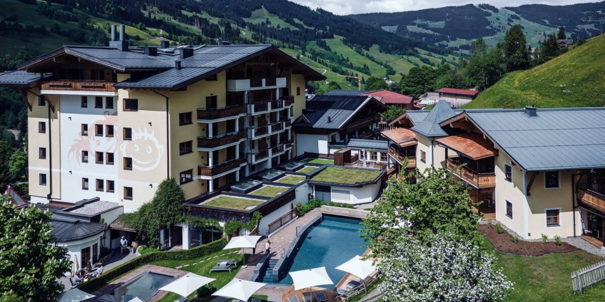 Ein mehrstöckiger Hotelkomplex mit einem zentralen Außenpool, Liegestühlen und Sonnenschirmen, umgeben von grünen Hügeln und Bergen unter einem bewölkten Himmel.