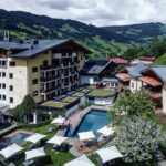 Ein mehrstöckiger Hotelkomplex mit einem zentralen Außenpool, Liegestühlen und Sonnenschirmen, umgeben von grünen Hügeln und Bergen unter einem bewölkten Himmel.
