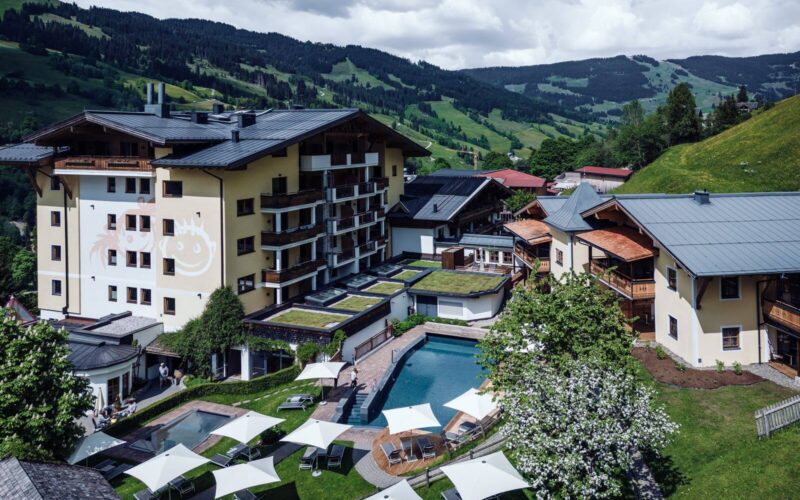 Ein mehrstöckiger Hotelkomplex mit einem zentralen Außenpool, Liegestühlen und Sonnenschirmen, umgeben von grünen Hügeln und Bergen unter einem bewölkten Himmel.