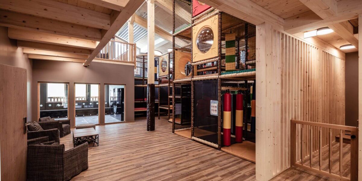 Geräumiger Indoor-Spielbereich mit Holzböden und -decken, einem Klettergerüst mit Netzen und Rutschen sowie einem Sitzbereich mit zwei Stühlen und einem kleinen Tisch.