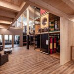 Geräumiger Indoor-Spielbereich mit Holzböden und -decken, einem Klettergerüst mit Netzen und Rutschen sowie einem Sitzbereich mit zwei Stühlen und einem kleinen Tisch.