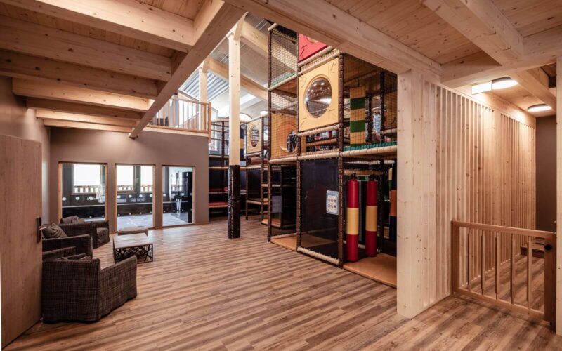 Geräumiger Indoor-Spielbereich mit Holzböden und -decken, einem Klettergerüst mit Netzen und Rutschen sowie einem Sitzbereich mit zwei Stühlen und einem kleinen Tisch.