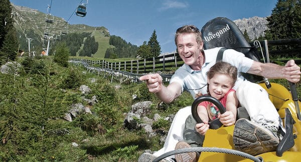 Ein Mann und ein junges Mädchen fahren mit einem gelben Alpine Coaster einen grasbewachsenen Berghang hinunter, mit Sesselliften und Kiefern im Hintergrund.