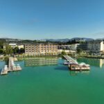 Ein Hotelkomplex am See mit drei markanten Gebäuden, Stegen mit Liegestühlen und klarem türkisfarbenem Wasser unter blauem Himmel.