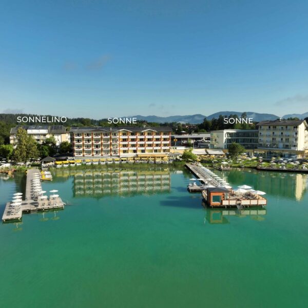 Ein Hotelkomplex am See mit drei markanten Gebäuden, Stegen mit Liegestühlen und klarem türkisfarbenem Wasser unter blauem Himmel.