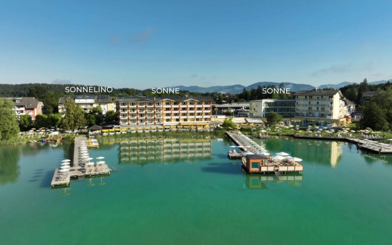 Ein Hotelkomplex am See mit drei markanten Gebäuden, Stegen mit Liegestühlen und klarem türkisfarbenem Wasser unter blauem Himmel.