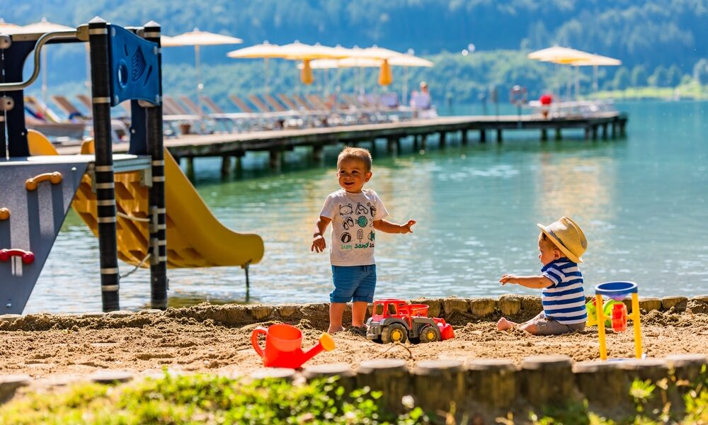 Zwei kleine Kinder spielen auf einer Sandfläche an einem See, auf der Spielzeug verstreut ist. Im Hintergrund sind eine Spielplatzrutsche und ein Steg mit Liegestühlen und Sonnenschirmen zu sehen.