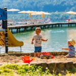 Zwei kleine Kinder spielen auf einer Sandfläche an einem See, auf der Spielzeug verstreut ist. Im Hintergrund sind eine Spielplatzrutsche und ein Steg mit Liegestühlen und Sonnenschirmen zu sehen.