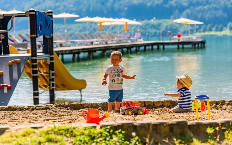 Zwei kleine Kinder spielen auf einer Sandfläche an einem See, auf der Spielzeug verstreut ist. Im Hintergrund sind eine Spielplatzrutsche und ein Steg mit Liegestühlen und Sonnenschirmen zu sehen.