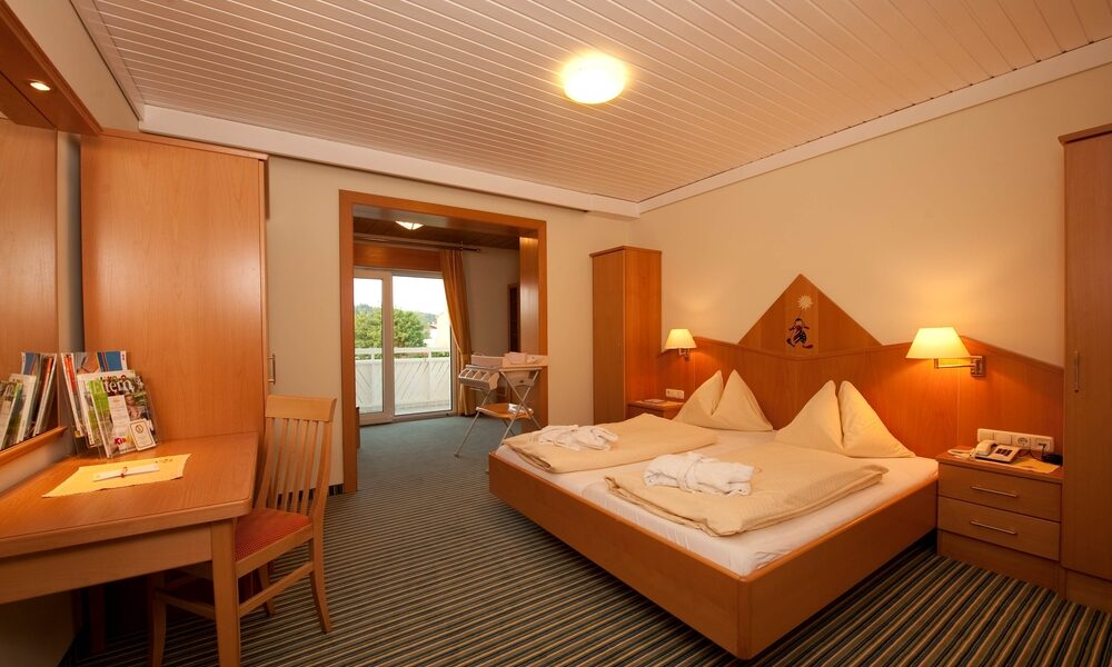 Ein Hotelzimmer mit einem Doppelbett, Nachttischen, Lampen, einem Schreibtisch mit einem Stuhl, Holzmöbeln und einem Blick durch eine offene Tür auf einen Balkon.