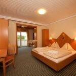 Ein Hotelzimmer mit einem Doppelbett, Nachttischen, Lampen, einem Schreibtisch mit einem Stuhl, Holzmöbeln und einem Blick durch eine offene Tür auf einen Balkon.