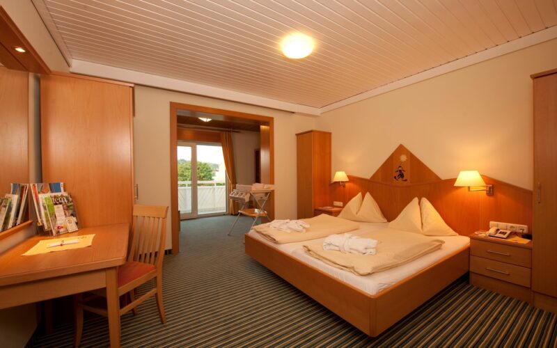 Ein Hotelzimmer mit einem Doppelbett, Nachttischen, Lampen, einem Schreibtisch mit einem Stuhl, Holzmöbeln und einem Blick durch eine offene Tür auf einen Balkon.