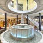 Ein blau beleuchteter Marmorbrunnen steht in der Mitte eines runden Raums mit Säulen und einer geschwungenen Treppe, die zu einem oberen Balkon führt.