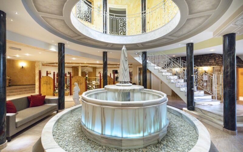 Ein blau beleuchteter Marmorbrunnen steht in der Mitte eines runden Raums mit Säulen und einer geschwungenen Treppe, die zu einem oberen Balkon führt.