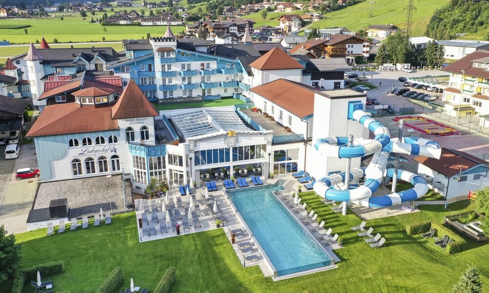 Luftaufnahme eines Resorts mit roten Dächern, Außenpool, Sonnenliegen und einer großen blau-weißen Wasserrutsche, umgeben von grünen Rasenflächen und nahe gelegenen Bergen.