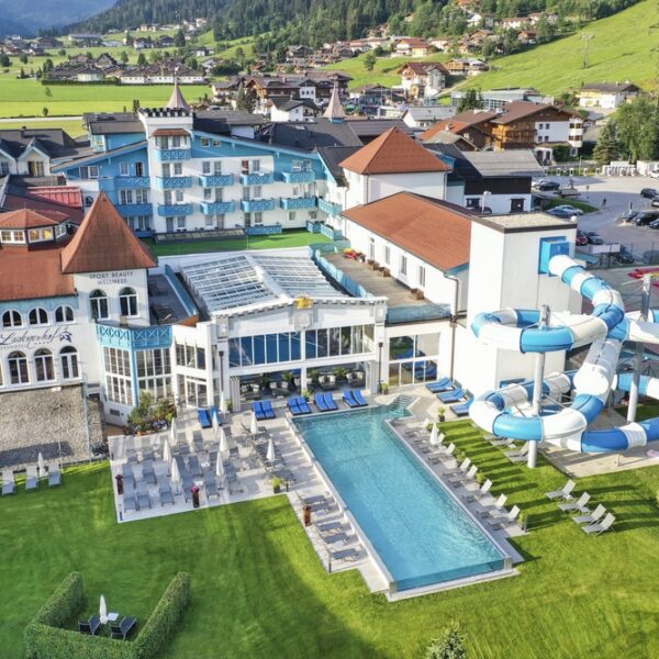 Luftaufnahme eines Resorts mit roten Dächern, Außenpool, Sonnenliegen und einer großen blau-weißen Wasserrutsche, umgeben von grünen Rasenflächen und nahe gelegenen Bergen.