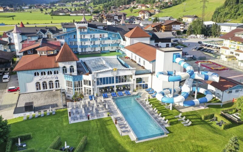 Luftaufnahme eines Resorts mit roten Dächern, Außenpool, Sonnenliegen und einer großen blau-weißen Wasserrutsche, umgeben von grünen Rasenflächen und nahe gelegenen Bergen.