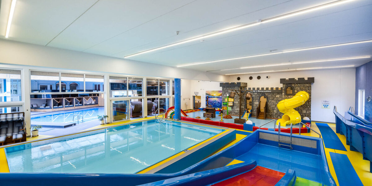 Innenbereich des Kinderbeckens mit bunten Wasserrutschen, einem flachen Becken und einer Spielstruktur in Form einer Burg mit einer gelben Spiralrutsche. Große Fenster geben den Blick auf einen weiteren Poolbereich frei.