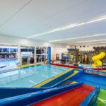 Innenbereich des Kinderbeckens mit bunten Wasserrutschen, einem flachen Becken und einer Spielstruktur in Form einer Burg mit einer gelben Spiralrutsche. Große Fenster geben den Blick auf einen weiteren Poolbereich frei.