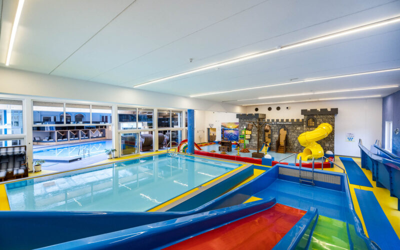 Innenbereich des Kinderbeckens mit bunten Wasserrutschen, einem flachen Becken und einer Spielstruktur in Form einer Burg mit einer gelben Spiralrutsche. Große Fenster geben den Blick auf einen weiteren Poolbereich frei.