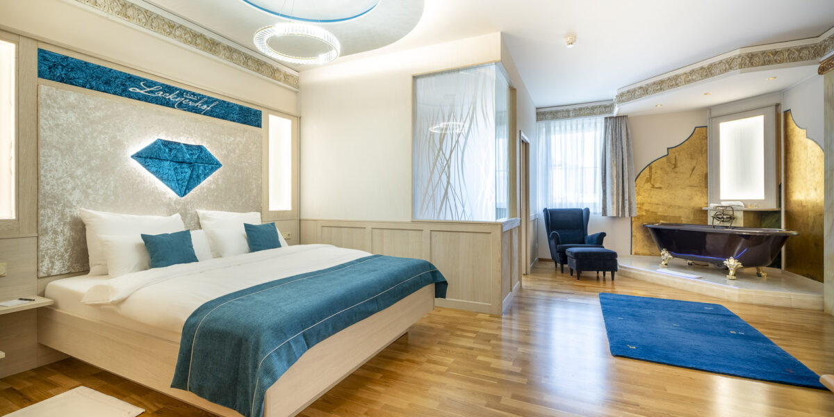 Modernes Hotelzimmer mit Doppelbett, blauen Akzenten, Sessel, goldgerahmter Badewanne, Holzfußboden und natürlichem Licht durch große Fenster.