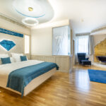 Modernes Hotelzimmer mit Doppelbett, blauen Akzenten, Sessel, goldgerahmter Badewanne, Holzfußboden und natürlichem Licht durch große Fenster.