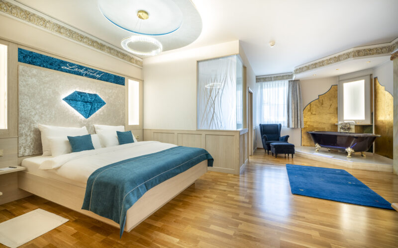 Modernes Hotelzimmer mit Doppelbett, blauen Akzenten, Sessel, goldgerahmter Badewanne, Holzfußboden und natürlichem Licht durch große Fenster.