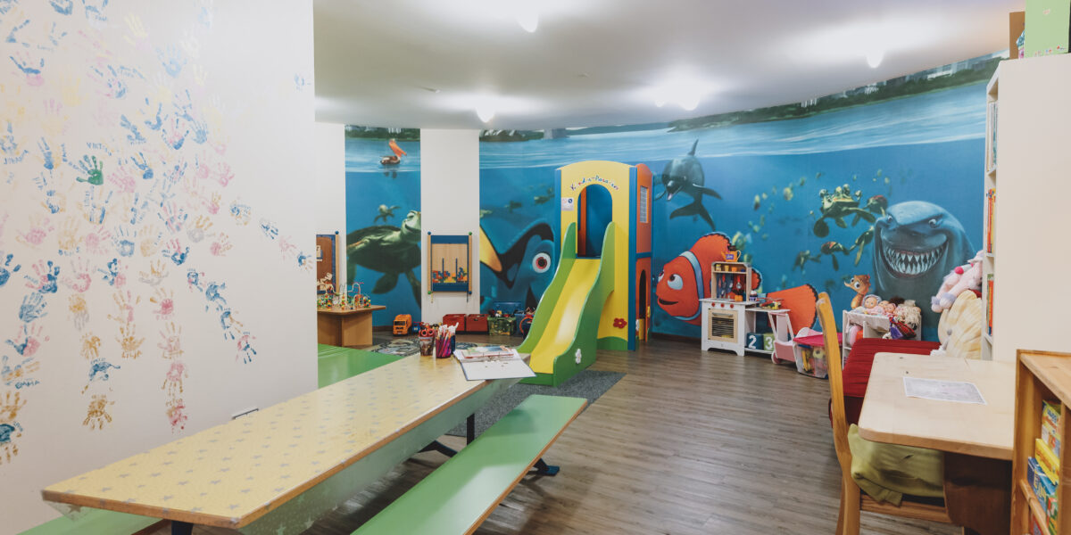 Kinderspielzimmer mit einer Wandmalerei von Findet Nemo, einer Rutsche, Spielzeug, Büchern, Tischen, Stühlen und bunten Handabdrücken an einer Wand.