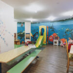 Kinderspielzimmer mit einer Wandmalerei von Findet Nemo, einer Rutsche, Spielzeug, Büchern, Tischen, Stühlen und bunten Handabdrücken an einer Wand.