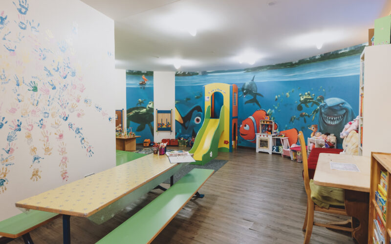 Kinderspielzimmer mit einer Wandmalerei von Findet Nemo, einer Rutsche, Spielzeug, Büchern, Tischen, Stühlen und bunten Handabdrücken an einer Wand.