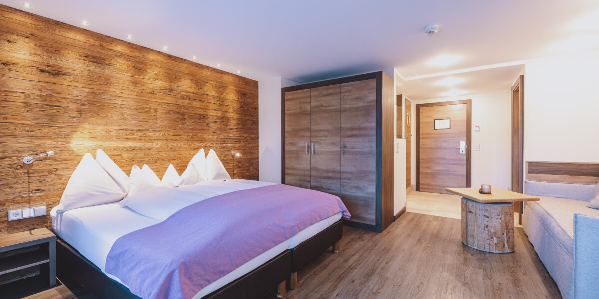 Modernes Hotelzimmer mit Doppelbett, Akzentwand aus Holz, Kleiderschrank, kleinem Tisch, Sofa und Holzfußboden, gut beleuchtet mit natürlichem und künstlichem Licht.