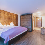 Modernes Hotelzimmer mit Doppelbett, Akzentwand aus Holz, Kleiderschrank, kleinem Tisch, Sofa und Holzfußboden, gut beleuchtet mit natürlichem und künstlichem Licht.