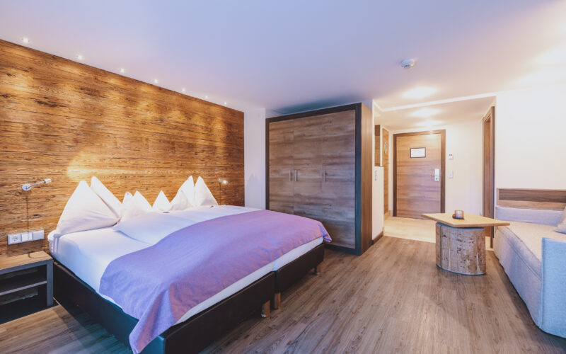 Modernes Hotelzimmer mit Doppelbett, Akzentwand aus Holz, Kleiderschrank, kleinem Tisch, Sofa und Holzfußboden, gut beleuchtet mit natürlichem und künstlichem Licht.