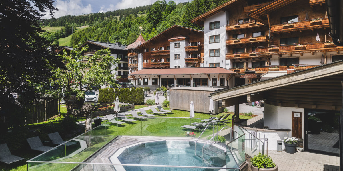 Ein Blick auf ein Hotel mit Holzbalkonen, umgeben von Grün und Bergen, mit einem Außenpool und Liegestühlen an einem sonnigen Tag.