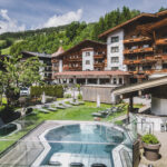 Ein Blick auf ein Hotel mit Holzbalkonen, umgeben von Grün und Bergen, mit einem Außenpool und Liegestühlen an einem sonnigen Tag.