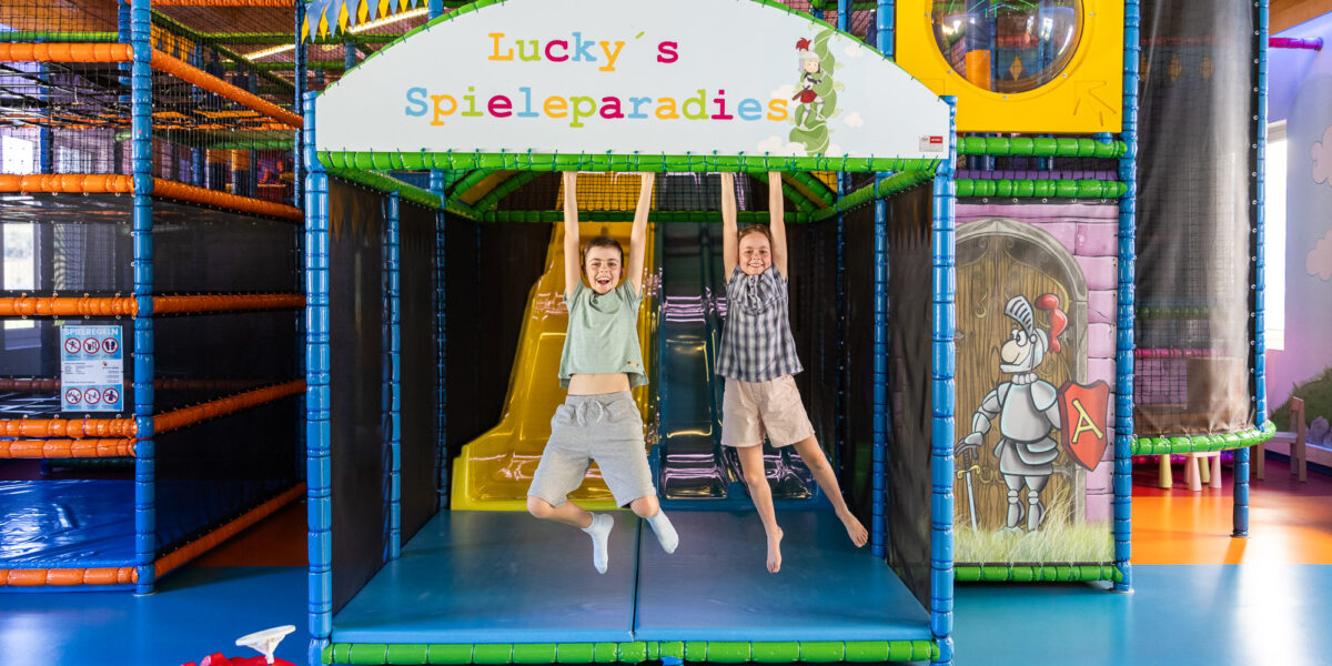 Zwei Kinder hängen an Stangen unter einem Schild mit der Aufschrift "Lucky's Spieleparadies" in einem farbenfrohen Indoor-Spielplatz, mit Rutschen und Spielgeräten im Hintergrund.