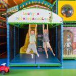 Zwei Kinder hängen an Stangen unter einem Schild mit der Aufschrift "Lucky's Spieleparadies" in einem farbenfrohen Indoor-Spielplatz, mit Rutschen und Spielgeräten im Hintergrund.