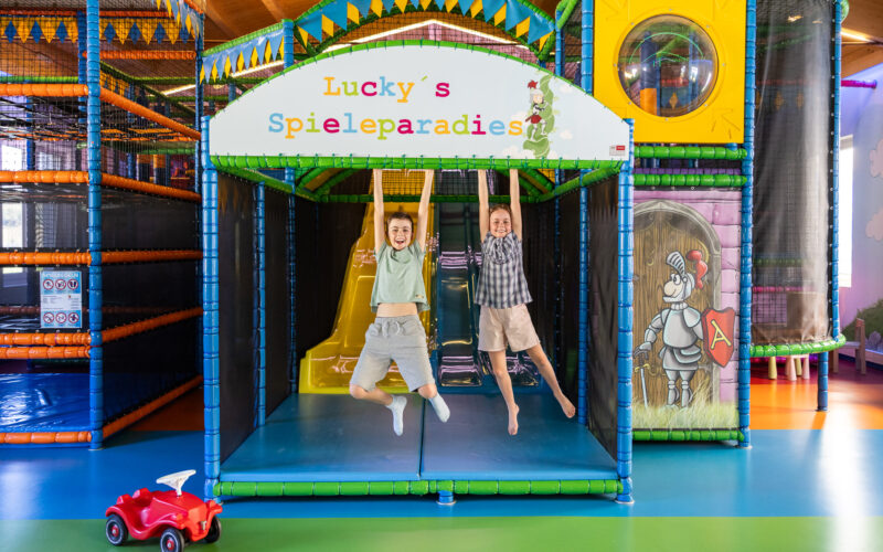 Zwei Kinder hängen an Stangen unter einem Schild mit der Aufschrift "Lucky's Spieleparadies" in einem farbenfrohen Indoor-Spielplatz, mit Rutschen und Spielgeräten im Hintergrund.