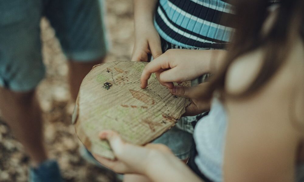 Zwei Kinder betrachten eine handgezeichnete Karte auf einem Stück Holz, während sie im Freien stehen.