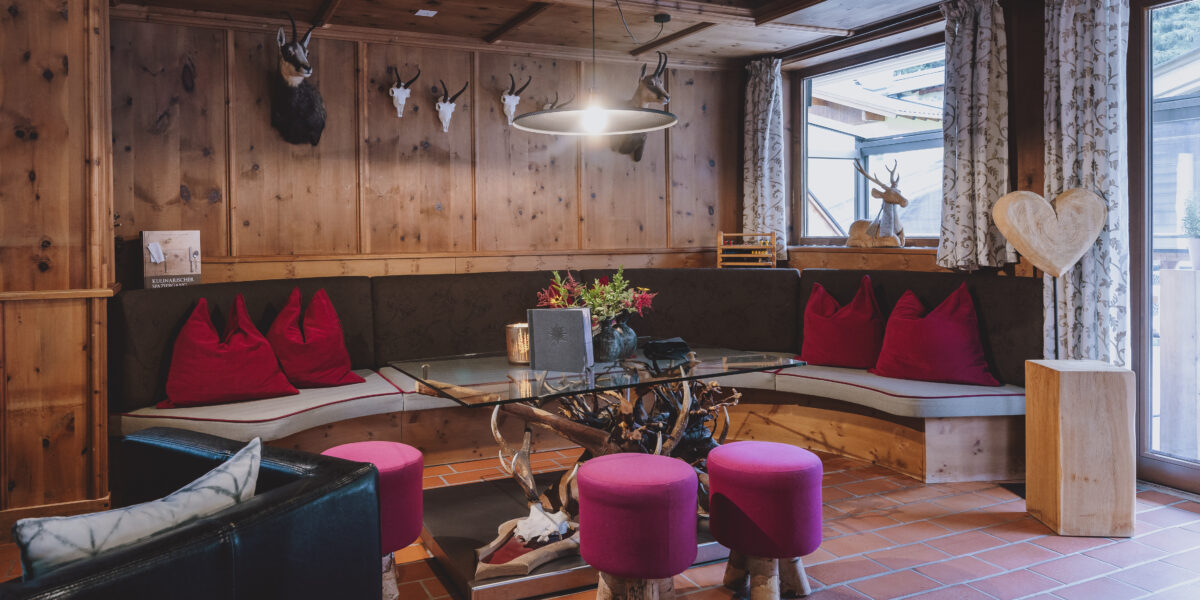Gemütliche Sitzecke aus Holz mit einer geschwungenen Bank, roten Kissen, runden Hockern, einem gläsernen Couchtisch, Geweihdekor und natürlichem Licht durch ein großes Fenster.