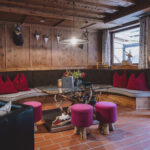 Gemütliche Sitzecke aus Holz mit einer geschwungenen Bank, roten Kissen, runden Hockern, einem gläsernen Couchtisch, Geweihdekor und natürlichem Licht durch ein großes Fenster.